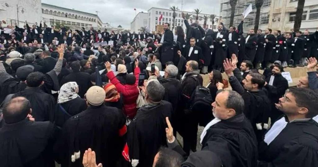 Réforme des avocats au Maroc : entre volonté de modernisation et lignes rouges de la profession Une foule importante d’avocats vêtus de robes noires se rassemble en plein air, probablement devant un bâtiment institutionnel. Certains sont debout sur des marches ou une estrade, prenant la parole ou levant les mains, tandis que d’autres, en contrebas, écoutent et participent en levant les bras. L’atmosphère évoque une mobilisation ou une manifestation collective de la profession.