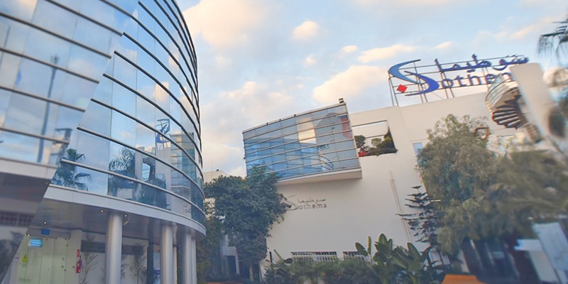 Façade moderne du siège de Sothema, composée de bâtiments aux murs vitrés et lignes courbes, entourée de végétation, sous un ciel légèrement nuageux.