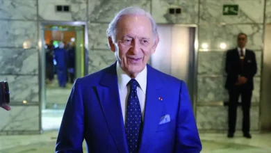 Homme âgé en costume bleu élégant et cravate assortie, portant une pochette et une décoration à la boutonnière, se tenant dans un hall en marbre éclairé, avec des agents de sécurité en arrière-plan près d’ascenseurs ouverts.