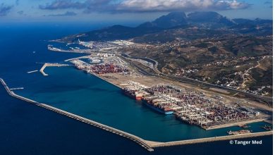 Vue aérienne d’un grand port industriel en bord de mer, avec de nombreux conteneurs empilés, des grues portuaires, plusieurs quais et digues protégeant le bassin. À l’arrière-plan, on distingue une ville étendue et des reliefs montagneux sous un ciel partiellement nuageux.