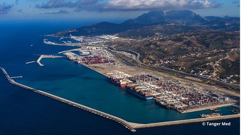 Vue aérienne d’un grand port industriel en bord de mer, avec de nombreux conteneurs empilés, des grues portuaires, plusieurs quais et digues protégeant le bassin. À l’arrière-plan, on distingue une ville étendue et des reliefs montagneux sous un ciel partiellement nuageux.