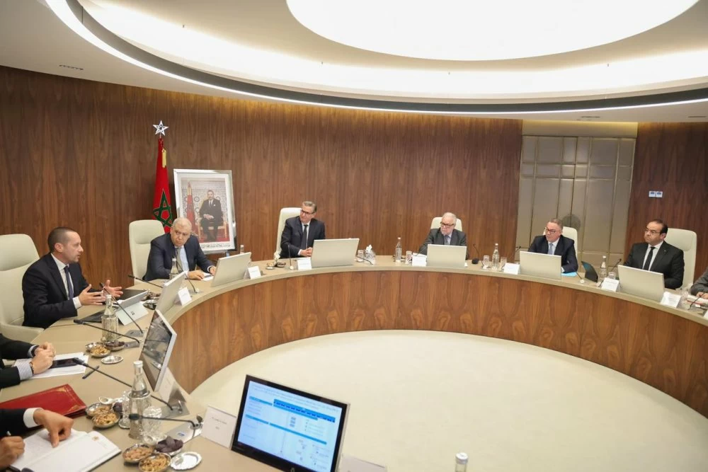 Réunion présidée par Aziz Akhannouch à Rabat sur la réforme du système de santé, avec des responsables autour d’une table de pilotage et le drapeau marocain en arrière-plan
