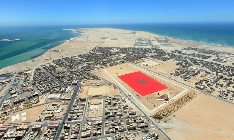 Vue aérienne de Dakhla montrant la ville entre lagune et océan Atlantique, avec une grande place arborant le drapeau marocain au cœur d’un environnement désertique.