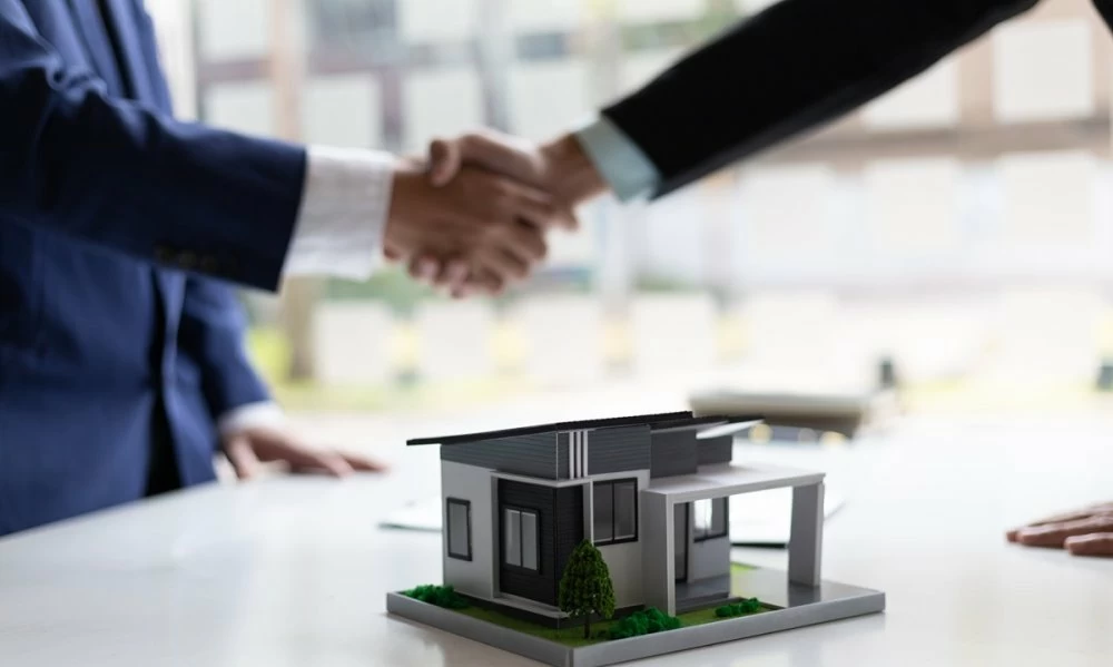 Poignée de main entre deux hommes en costume scellant un accord immobilier, avec une maquette de maison posée sur une table au premier plan, symbolisant une transaction ou un partenariat dans le secteur de l’immobilier.
