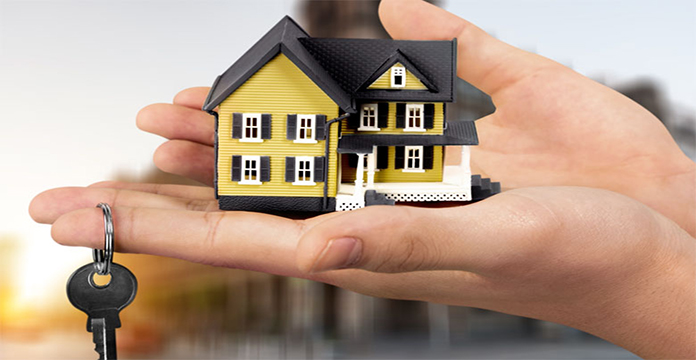 Une main tient une petite maquette de maison jaune avec un toit noir, tandis qu’une clé est suspendue à un porte-clés à proximité, symbolisant l’achat ou la location d’un bien immobilier.