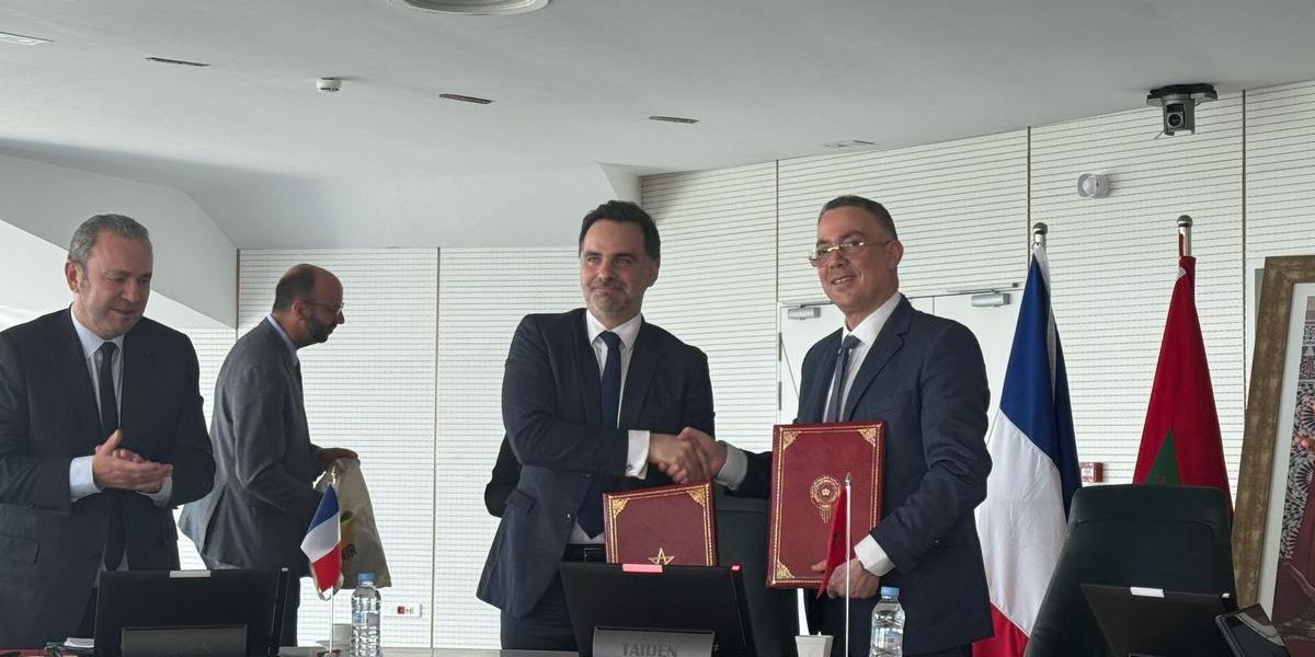 Une scène officielle de signature d’accord dans une salle de réunion moderne. Au centre, deux hommes en costume se serrent la main en souriant tout en tenant des dossiers rouges ornés d’un emblème, symbolisant un partenariat. Derrière eux, les drapeaux du Maroc et de la France sont visibles, suggérant une coopération bilatérale. À gauche, deux autres hommes en costume assistent à la scène, l’un applaudissant. Une table de conférence avec des microphones et des bouteilles d’eau complète le cadre institutionnel.