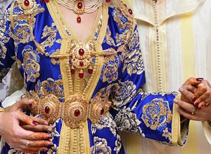 Une mariée portant un caftan bleu richement brodé d’or et ornée de bijoux traditionnels imposants, notamment une ceinture dorée (mdamma). Ses mains sont décorées au henné, et elle est accompagnée d’une autre personne vêtue de blanc, probablement lors d’une cérémonie de mariage traditionnelle marocaine.