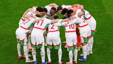 Joueurs de l’équipe nationale du Maroc regroupés en cercle sur la pelouse, en tenue blanche avec des touches rouges et vertes, en train de se motiver avant un match.