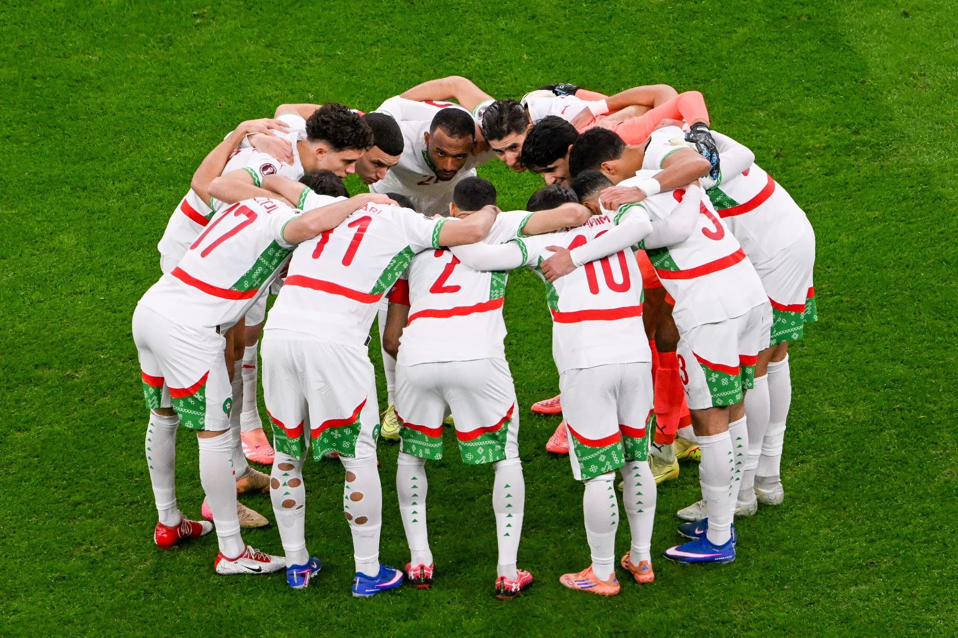 Joueurs de l’équipe nationale du Maroc regroupés en cercle sur la pelouse, en tenue blanche avec des touches rouges et vertes, en train de se motiver avant un match.