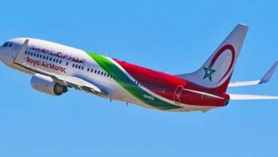 Un avion de la compagnie Royal Air Maroc en plein vol, avec une livrée blanche ornée de motifs rouges et verts inspirés du drapeau marocain. Le logo de la compagnie et l’inscription « Royal Air Maroc » sont visibles sur le fuselage, tandis que la dérive porte une étoile verte sur fond rouge. L’appareil évolue dans un ciel clair.