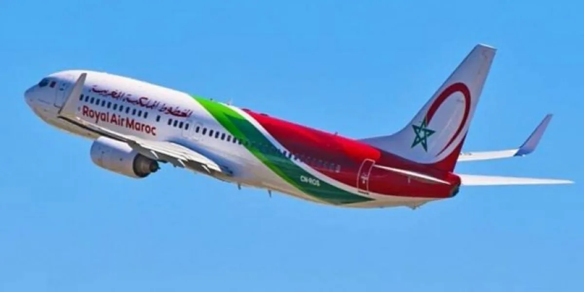 Un avion de la compagnie Royal Air Maroc en plein vol, avec une livrée blanche ornée de motifs rouges et verts inspirés du drapeau marocain. Le logo de la compagnie et l’inscription « Royal Air Maroc » sont visibles sur le fuselage, tandis que la dérive porte une étoile verte sur fond rouge. L’appareil évolue dans un ciel clair.