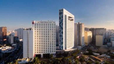 Vue urbaine en hauteur montrant un ensemble d’hôtels modernes au cœur d’une grande ville, dominé par un bâtiment blanc de grande hauteur avec une façade géométrique. À ses côtés, un hôtel Novotel et un ibis sont visibles, entourés d’immeubles, de routes animées et de palmiers, sous une lumière de fin de journée.