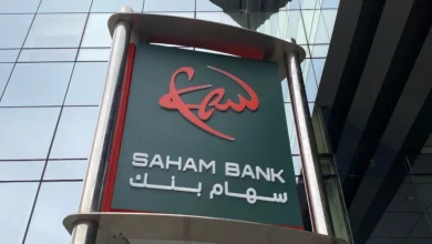 Façade d’un bâtiment moderne avec une enseigne de Saham Bank, affichant le logo rouge stylisé au-dessus du nom de la banque en lettres blanches, accompagné de son appellation en arabe « سهام بنك ». L’enseigne est fixée sur une structure métallique devant une paroi vitrée reflétant l’environnement urbain.