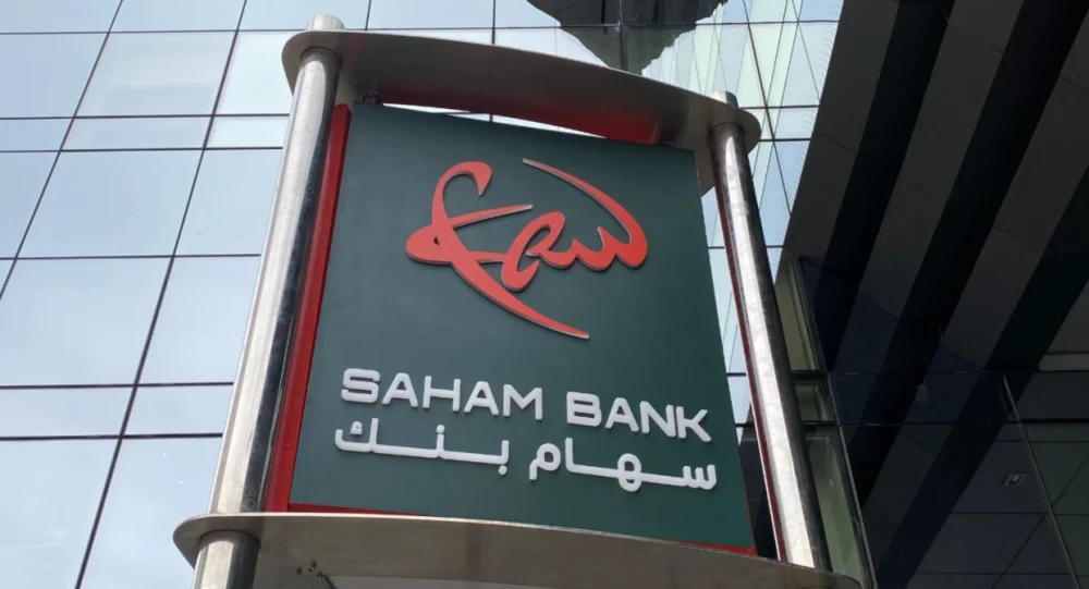 Façade d’un bâtiment moderne avec une enseigne de Saham Bank, affichant le logo rouge stylisé au-dessus du nom de la banque en lettres blanches, accompagné de son appellation en arabe « سهام بنك ». L’enseigne est fixée sur une structure métallique devant une paroi vitrée reflétant l’environnement urbain.