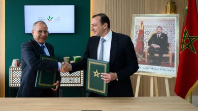 oignée de main entre deux responsables lors d’une cérémonie officielle de signature d’accords, chacun tenant un dossier vert orné de l’étoile marocaine. La scène se déroule dans un cadre institutionnel avec un écran affichant le logo du Crédit Agricole du Maroc en arrière-plan. À droite, un portrait du Roi Mohammed VI et un drapeau marocain renforcent le caractère officiel de l’événement.