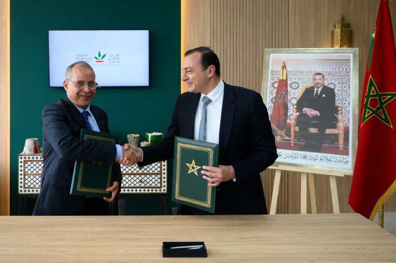 oignée de main entre deux responsables lors d’une cérémonie officielle de signature d’accords, chacun tenant un dossier vert orné de l’étoile marocaine. La scène se déroule dans un cadre institutionnel avec un écran affichant le logo du Crédit Agricole du Maroc en arrière-plan. À droite, un portrait du Roi Mohammed VI et un drapeau marocain renforcent le caractère officiel de l’événement.