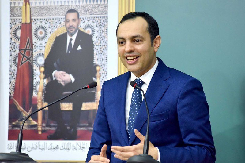 Responsable marocain s’exprimant lors d’une réunion officielle, assis devant un micro, avec le drapeau du Maroc et un portrait du Roi en arrière-plan