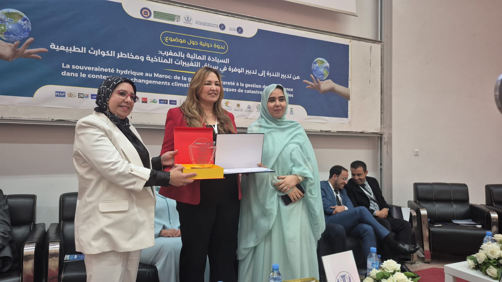 Trois femmes posent lors d’une cérémonie officielle en intérieur, en tenant un trophée et un certificat. Elles se tiennent devant une bannière dédiée à une conférence sur la souveraineté hydrique au Maroc et les enjeux liés aux changements climatiques. Des participants assis en arrière-plan assistent à l’événement, dans une ambiance formelle.
