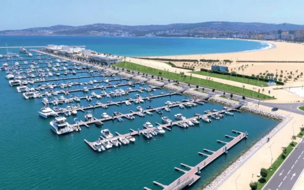 Vue aérienne d’une marina moderne au Maroc avec des bateaux amarrés, située en bord de mer près d’une plage aménagée, illustrant le développement des infrastructures touristiques et maritimes.