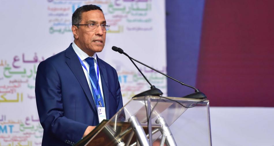 Un responsable syndical s’exprime lors d’une conférence, debout derrière un pupitre équipé de microphones. Vêtu d’un costume bleu et d’une cravate, il intervient dans un cadre officiel, avec un arrière-plan affichant des inscriptions liées à une organisation ou un événement, suggérant une prise de parole institutionnelle ou syndicale.