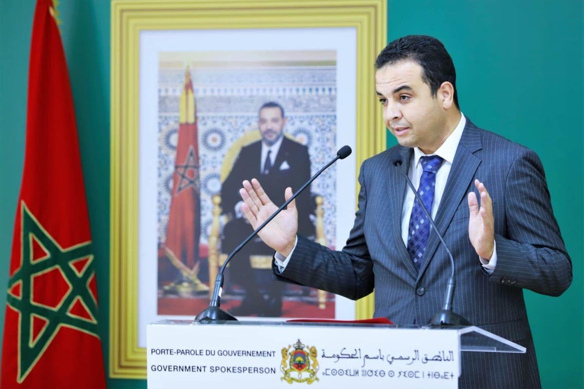 Un homme en costume sombre, debout derrière un pupitre officiel, s’exprime lors d’une conférence de presse. Il gesticule avec les mains pour appuyer son discours. Derrière lui, on distingue le drapeau marocain ainsi qu’un portrait du roi encadré, dans un décor institutionnel.