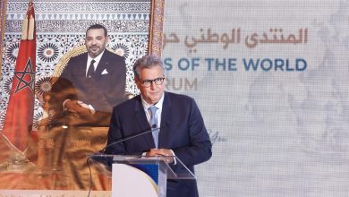 Un homme en costume prend la parole derrière un pupitre lors d’un événement officiel. À côté de lui se trouve un grand portrait du roi du Maroc devant le drapeau marocain. L’arrière-plan affiche partiellement du texte en arabe et en anglais lié à un forum international.