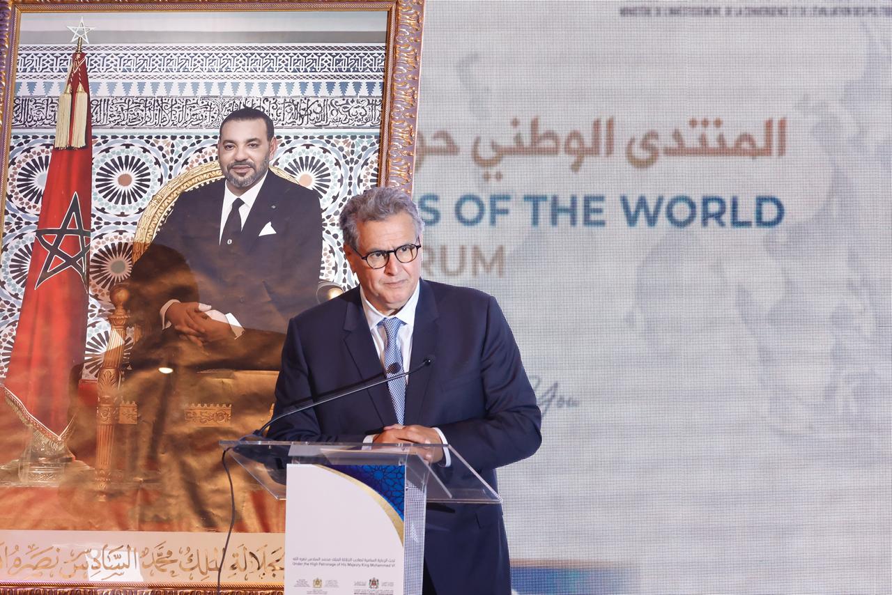 Un homme en costume prend la parole derrière un pupitre lors d’un événement officiel. À côté de lui se trouve un grand portrait du roi du Maroc devant le drapeau marocain. L’arrière-plan affiche partiellement du texte en arabe et en anglais lié à un forum international.