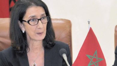 Une femme portant des lunettes s’exprime lors d’une réunion officielle, assise devant un microphone avec un drapeau marocain posé sur la table.