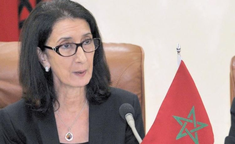 Une femme portant des lunettes s’exprime lors d’une réunion officielle, assise devant un microphone avec un drapeau marocain posé sur la table.