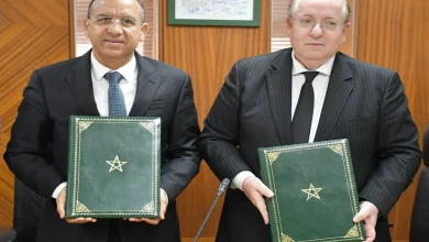 Deux responsables en costume officiel posent dans une salle de réunion après une cérémonie de signature, chacun tenant un dossier vert orné de l’emblème du Royaume du Maroc. En arrière-plan, des panneaux en bois et un document encadré sont visibles, traduisant un cadre institutionnel et formel.