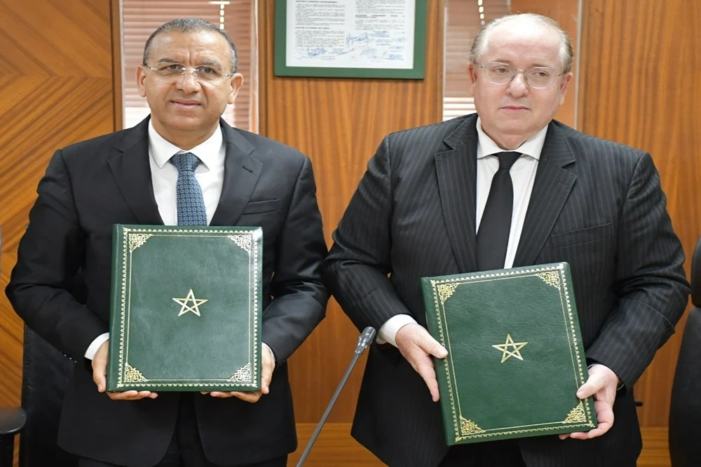 Deux responsables en costume officiel posent dans une salle de réunion après une cérémonie de signature, chacun tenant un dossier vert orné de l’emblème du Royaume du Maroc. En arrière-plan, des panneaux en bois et un document encadré sont visibles, traduisant un cadre institutionnel et formel.