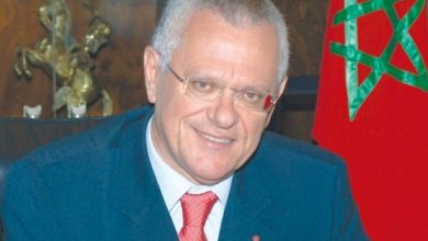 Driss Benhima en costume et cravate rouge, souriant devant un drapeau marocain dans un bureau officiel.