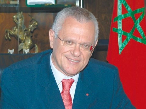 Driss Benhima en costume et cravate rouge, souriant devant un drapeau marocain dans un bureau officiel.