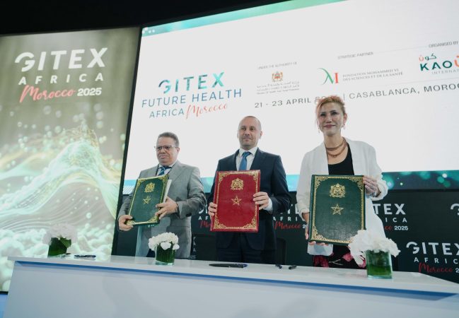 Signature officielle lors de GITEX Future Health Africa à Casablanca, avec trois responsables présentant des dossiers après un partenariat, devant un écran affichant l’événement.