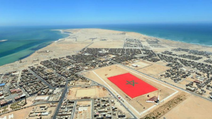 Vue aérienne d’une ville du Sahara marocain bordée par l’océan, avec un immense drapeau marocain peint au sol au centre d’un vaste espace désertique.