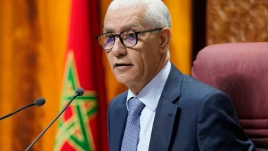 Homme en costume et lunettes prenant la parole lors d’une séance officielle, assis devant un micro avec le drapeau marocain en arrière-plan.