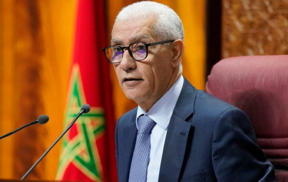 Homme en costume et lunettes prenant la parole lors d’une séance officielle, assis devant un micro avec le drapeau marocain en arrière-plan.