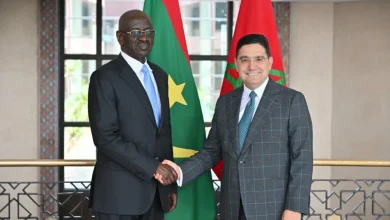Nasser Bourita Accueille le Ministre Mauritanien des Affaires Étrangères à Rabat 3 Deux hommes en costume se serrent la main devant les drapeaux du Maroc et d’un pays africain. Ils posent dans un bâtiment officiel lumineux, avec un décor sobre et élégant en arrière-plan.