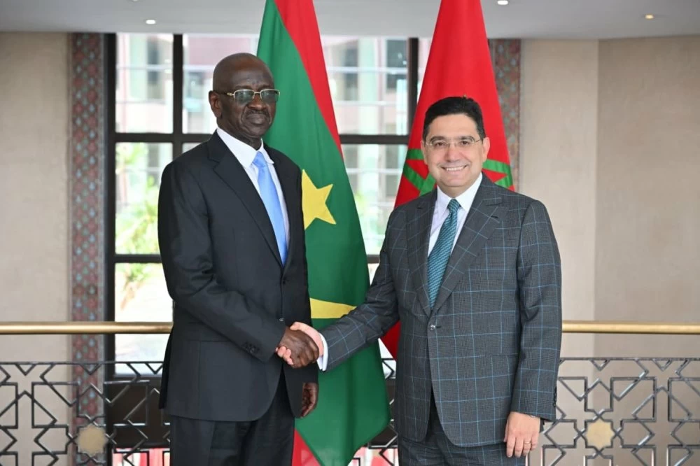Deux hommes en costume se serrent la main devant les drapeaux du Maroc et d’un pays africain. Ils posent dans un bâtiment officiel lumineux, avec un décor sobre et élégant en arrière-plan.