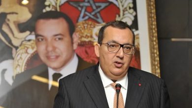 Portrait d’Ali Fassi Fihri lors d’une intervention officielle, vêtu d’un costume sombre et d’une cravate claire, s’exprimant derrière un microphone. En arrière-plan, un portrait du Roi Mohammed VI et un drapeau marocain encadré sont visibles.