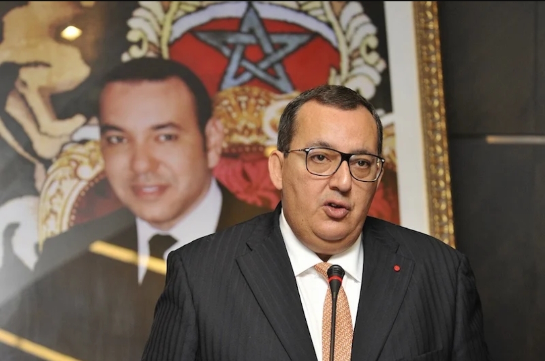 Décès d’Ali Fassi Fihri, ancien patron de l’ONEE et figure du management public marocain 1 Portrait d’Ali Fassi Fihri lors d’une intervention officielle, vêtu d’un costume sombre et d’une cravate claire, s’exprimant derrière un microphone. En arrière-plan, un portrait du Roi Mohammed VI et un drapeau marocain encadré sont visibles.