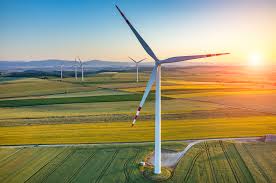 Paysage rural avec une éolienne au premier plan, entourée de champs agricoles aux couleurs variées, sous un ciel dégagé au coucher du soleil. D’autres éoliennes sont visibles à l’horizon, symbolisant la production d’énergie renouvelable.