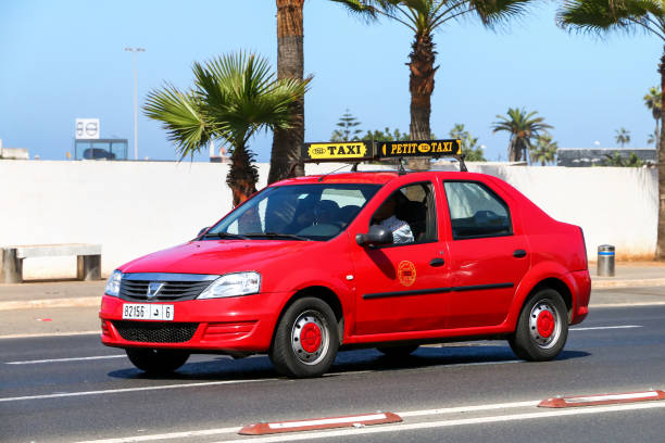 Un petit taxi rouge circule sur une avenue bordée de palmiers, sous un ciel dégagé. Le véhicule, de type berline, porte une enseigne « Petit Taxi » sur son toit et roule sur une large chaussée urbaine.