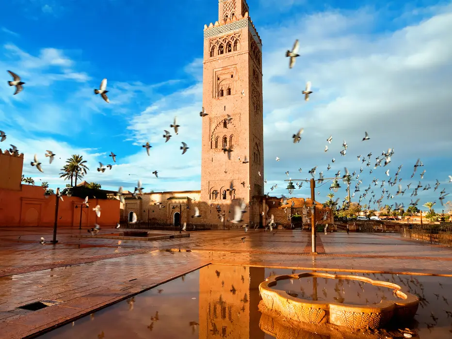 Vue de la Mosquée Koutoubia à Marrakech, avec son minaret emblématique dominant une place pavée encore humide après la pluie. Des pigeons s’envolent en groupe dans un ciel partiellement nuageux, tandis qu’un bassin en pierre au premier plan reflète la lumière dorée du soleil.