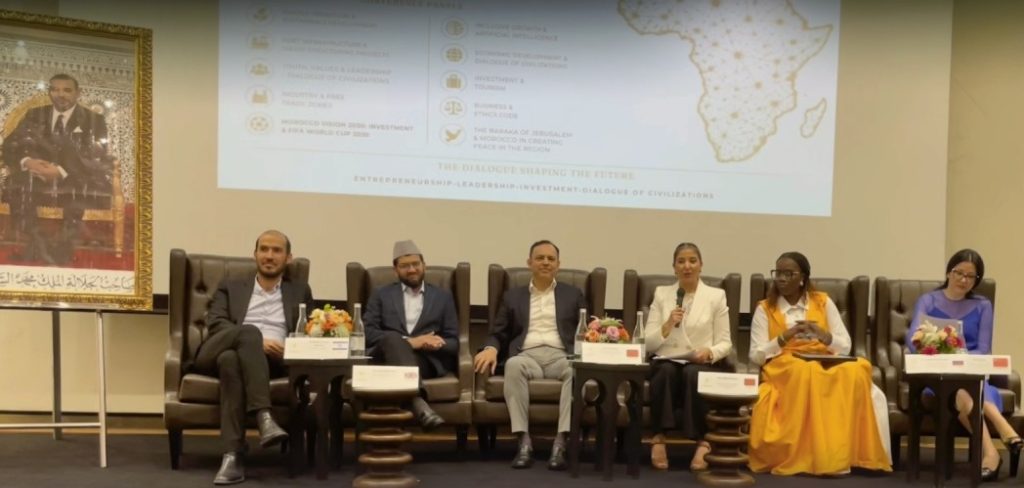 Tanger : Le Forum International « ELI Morocco 2026 » lie investissement, capital humain et coexistence Photographie d’un panel de discussion organisé dans une salle de conférence. Six intervenants sont assis sur de grands fauteuils en cuir sur une scène. Une femme au centre, vêtue d’un tailleur blanc, prend la parole avec un microphone à la main. Derrière eux, un écran de projection affiche les thèmes du forum liés à l’entrepreneuriat, au leadership, à l’investissement, à la croissance inclusive et au dialogue des civilisations, accompagnés d’une carte de l’Afrique. À gauche de la scène, un grand portrait officiel de Sa Majesté le Roi Mohammed VI est exposé. Des bouquets de fleurs décorent les tables placées devant les participants, dans une ambiance institutionnelle et internationale.