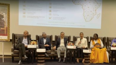 Tanger : Le Forum International « ELI Morocco 2026 » lie investissement, capital humain et coexistence 5 Photographie d’un panel de discussion organisé dans une salle de conférence. Six intervenants sont assis sur de grands fauteuils en cuir sur une scène. Une femme au centre, vêtue d’un tailleur blanc, prend la parole avec un microphone à la main. Derrière eux, un écran de projection affiche les thèmes du forum liés à l’entrepreneuriat, au leadership, à l’investissement, à la croissance inclusive et au dialogue des civilisations, accompagnés d’une carte de l’Afrique. À gauche de la scène, un grand portrait officiel de Sa Majesté le Roi Mohammed VI est exposé. Des bouquets de fleurs décorent les tables placées devant les participants, dans une ambiance institutionnelle et internationale.