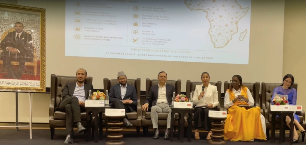 Photographie d’un panel de discussion organisé dans une salle de conférence. Six intervenants sont assis sur de grands fauteuils en cuir sur une scène. Une femme au centre, vêtue d’un tailleur blanc, prend la parole avec un microphone à la main. Derrière eux, un écran de projection affiche les thèmes du forum liés à l’entrepreneuriat, au leadership, à l’investissement, à la croissance inclusive et au dialogue des civilisations, accompagnés d’une carte de l’Afrique. À gauche de la scène, un grand portrait officiel de Sa Majesté le Roi Mohammed VI est exposé. Des bouquets de fleurs décorent les tables placées devant les participants, dans une ambiance institutionnelle et internationale.