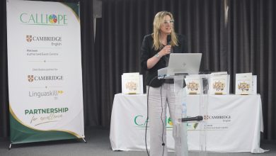 Une femme blonde portant des lunettes prend la parole derrière un pupitre transparent, micro à la main, lors d’une présentation organisée par Calliope. À sa gauche, une bannière affiche les logos de Calliope et Cambridge English, avec la mention « Platinum Authorised Exam Centre » et « Partnership for Excellence ». Un ordinateur portable est posé sur le pupitre, tandis que plusieurs exemplaires d’un ouvrage sont exposés sur une table recouverte d’une nappe blanche en arrière-plan. La scène se déroule dans une salle de conférence devant un rideau sombre.