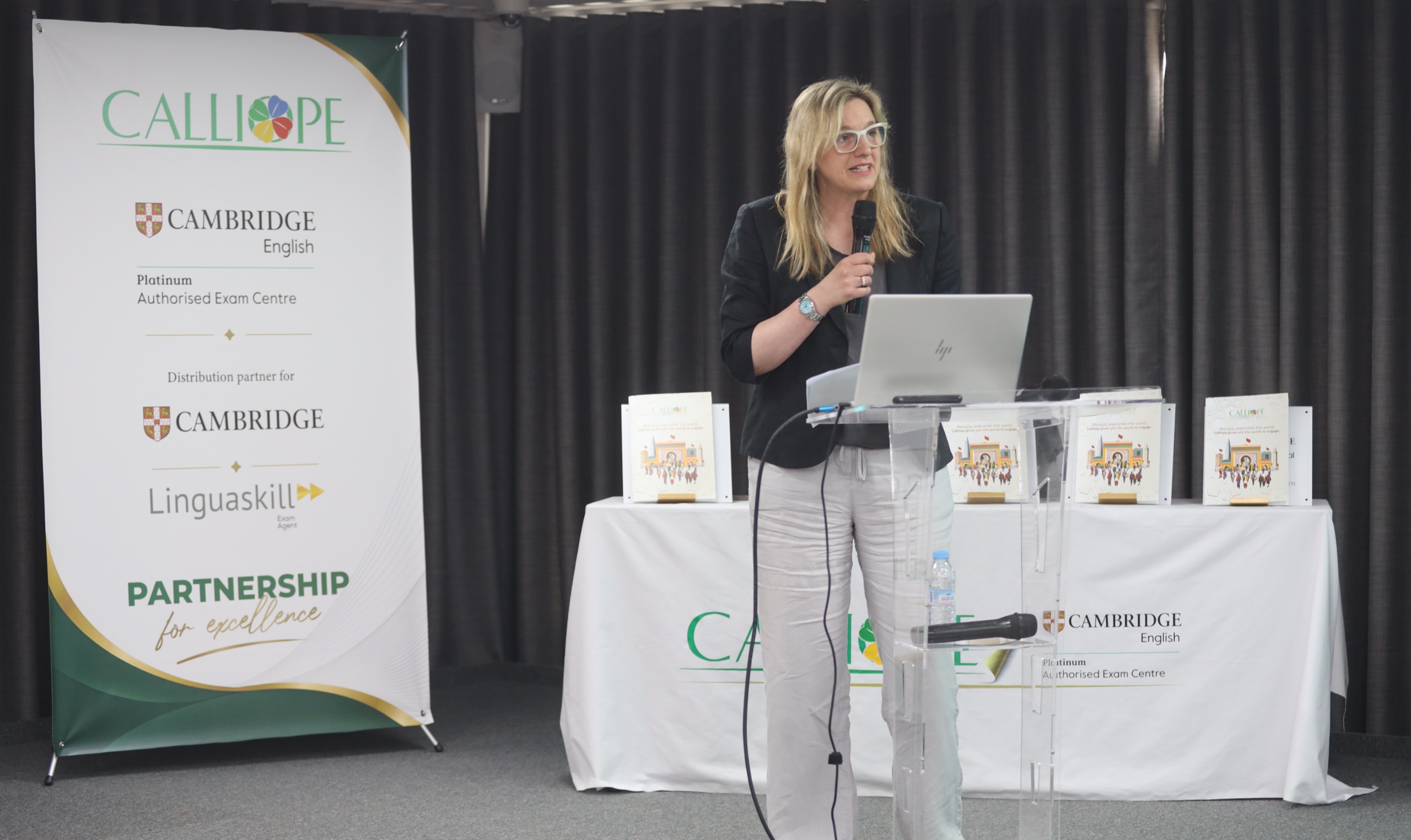 Une femme blonde portant des lunettes prend la parole derrière un pupitre transparent, micro à la main, lors d’une présentation organisée par Calliope. À sa gauche, une bannière affiche les logos de Calliope et Cambridge English, avec la mention « Platinum Authorised Exam Centre » et « Partnership for Excellence ». Un ordinateur portable est posé sur le pupitre, tandis que plusieurs exemplaires d’un ouvrage sont exposés sur une table recouverte d’une nappe blanche en arrière-plan. La scène se déroule dans une salle de conférence devant un rideau sombre.