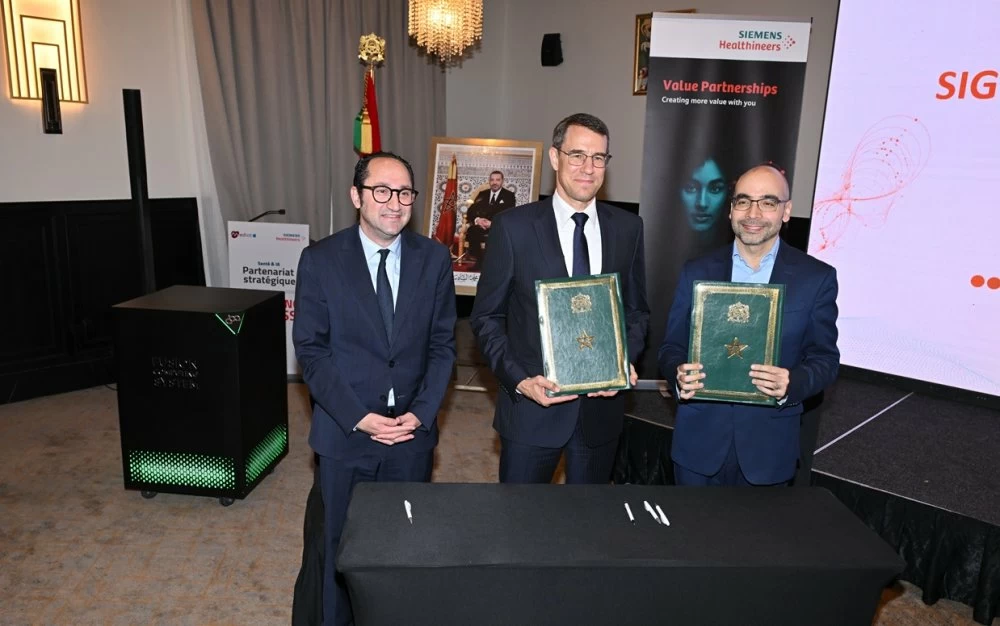 Signature d’un partenariat stratégique entre Siemens Healthineers et Mediot AI lors d’une cérémonie officielle au Maroc. Trois dirigeants en costume posent devant une table de signature, tandis que deux représentants tiennent les accords signés. À l’arrière-plan figurent le drapeau marocain, un portrait officiel du Roi Mohammed VI et les supports visuels de Siemens Healthineers, illustrant une alliance axée sur l’intelligence artificielle appliquée à la santé et aux infrastructures hospitalières en Afrique.